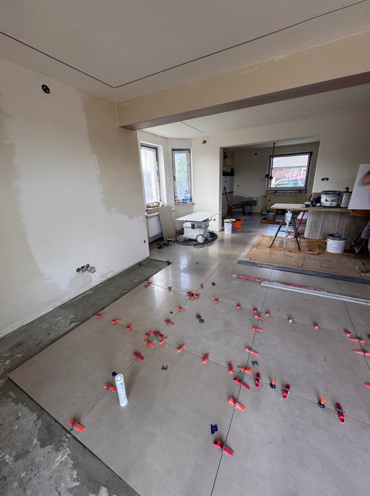 Układanie płytek podłogowych z użyciem klipsów poziomujących w przestronnym wnętrzu z oknami i przygotowaną ścianą. Widoczny sprzęt glazurniczy.