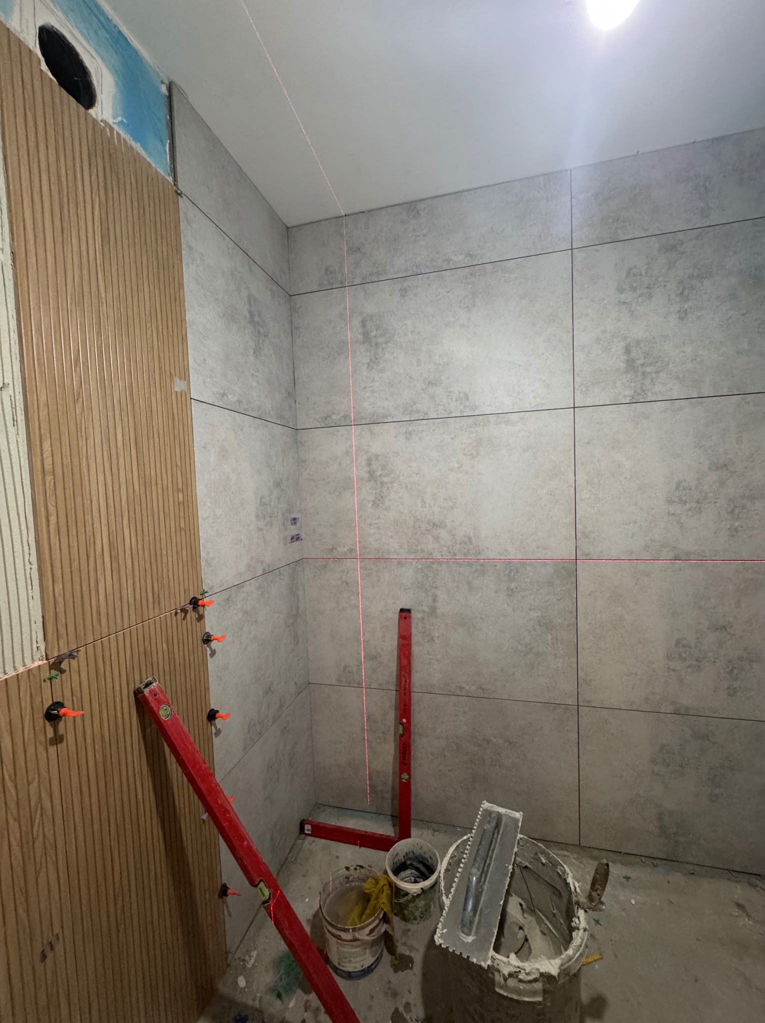 Montaż płytek imitujących drewno na ścianie łazienki, z użyciem poziomicy laserowej i klipsów. Częściowo wykończona ściana z szarych płytek.