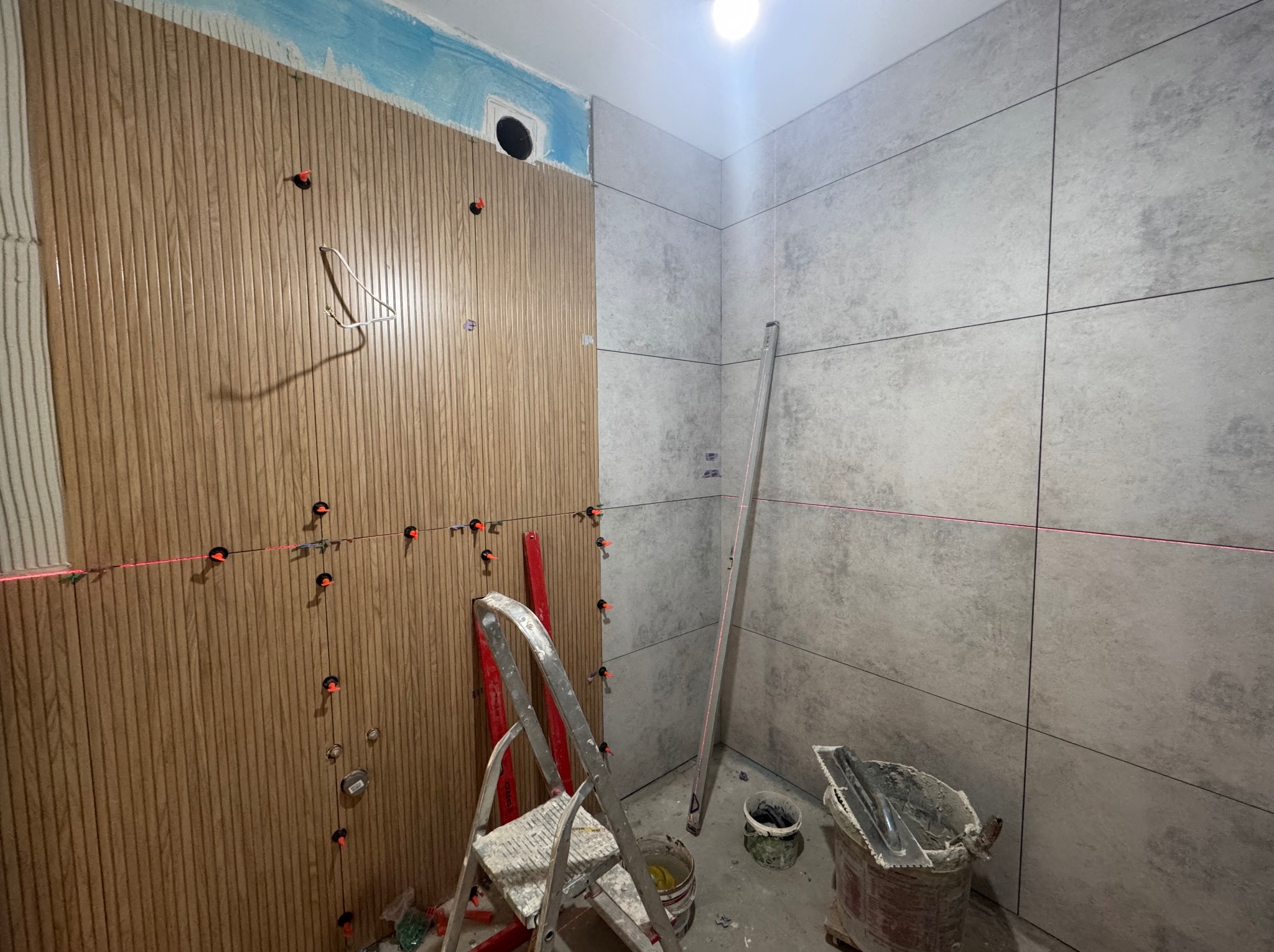 Montaż okładziny ściennej w łazience: płytki imitujące drewno i szare płytki, poziomica laserowa, drabina, etap prac wykończeniowych.