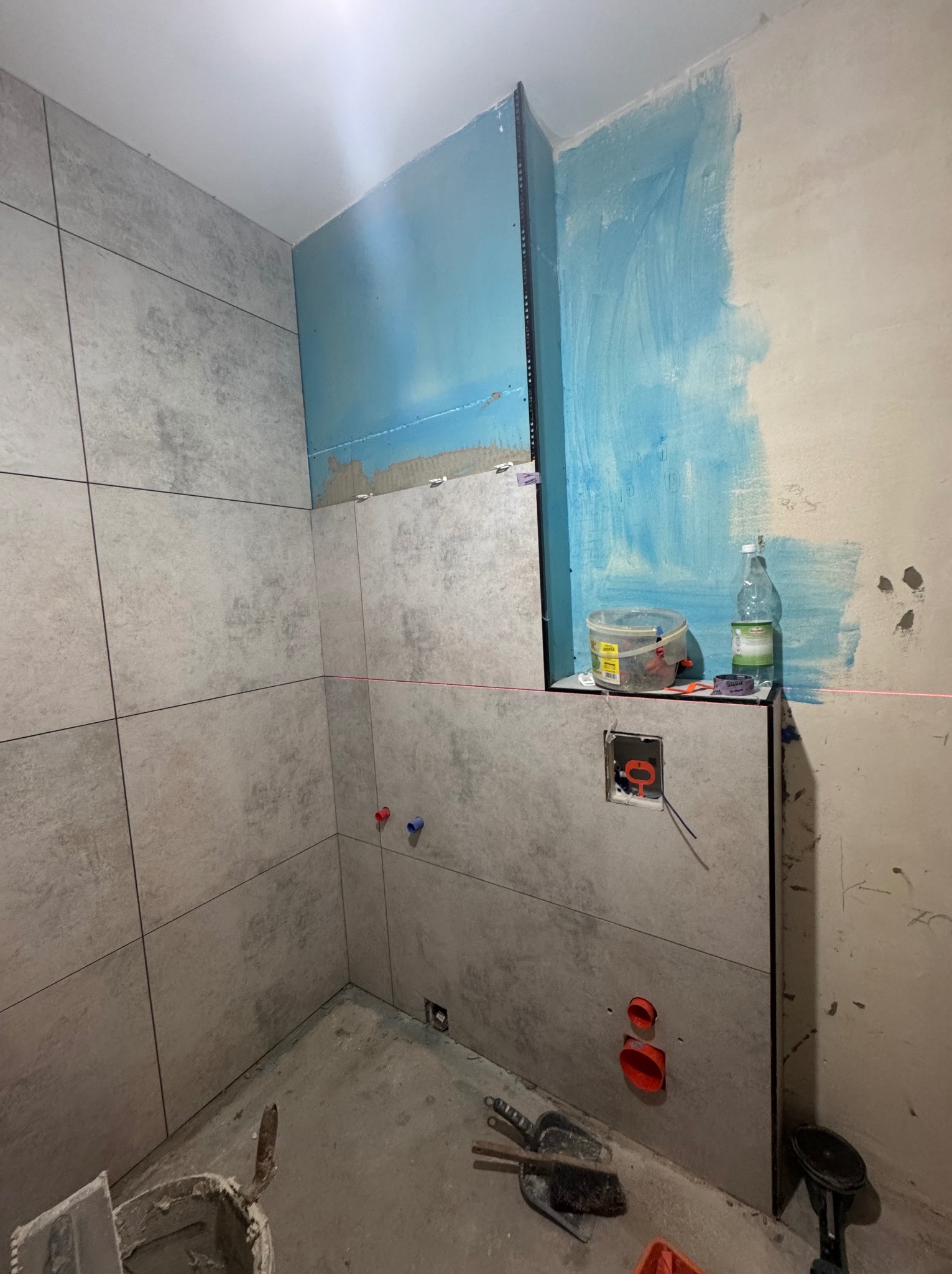 Wnętrze łazienki w trakcie wykańczania: szare płytki na ścianach, fragment ściany z niebieskim podkładem, poziomica laserowa, narzędzia glazurnicze na podłodze.