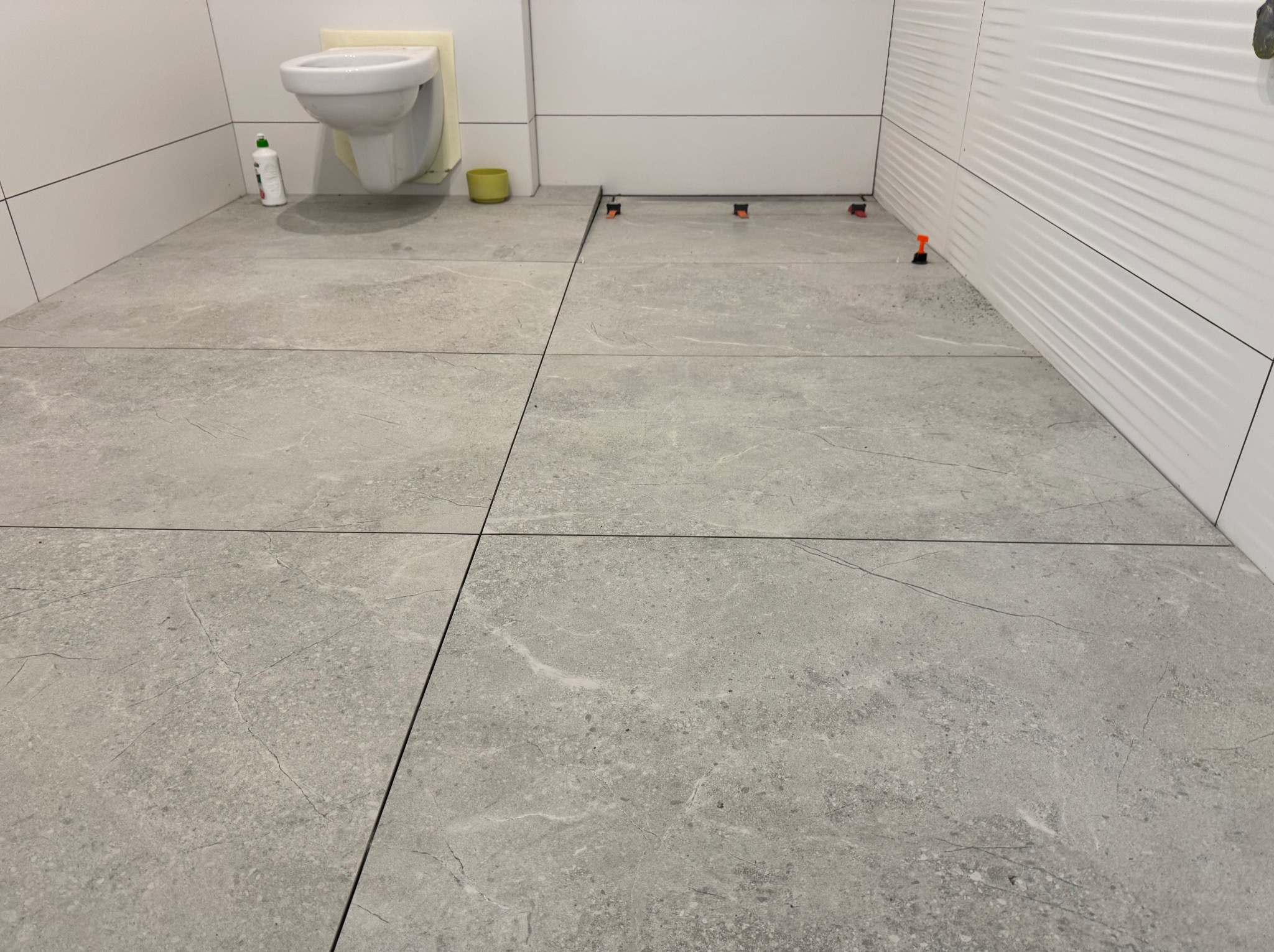 Szare płytki podłogowe w łazience, ułożone z użyciem systemu poziomowania. Widoczne płytki na ścianie i podwieszana toaleta z zabezpieczeniem.