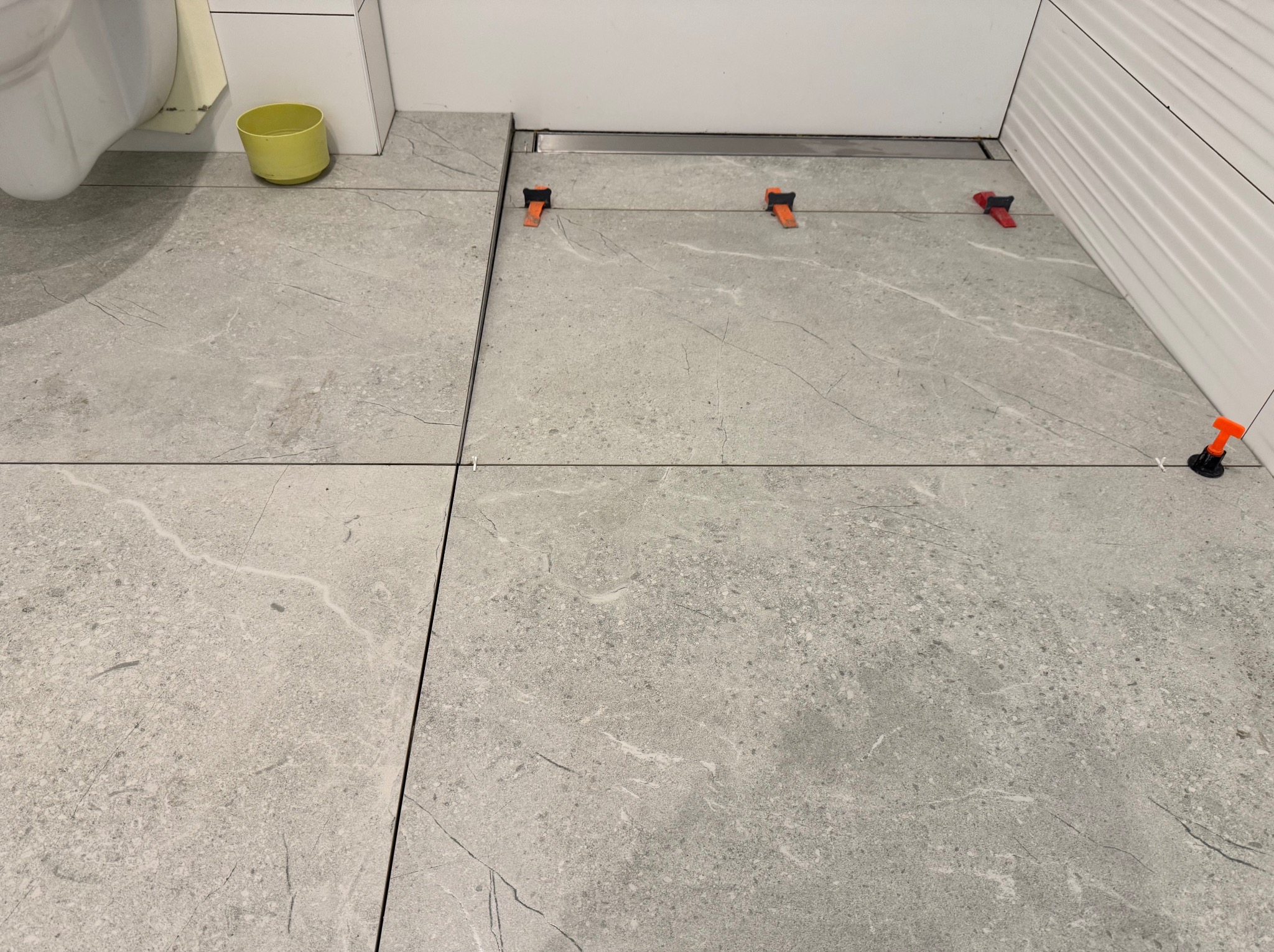 Szare płytki podłogowe w łazience, ułożone z użyciem klipsów poziomujących. Widoczny odpływ liniowy i fragment toalety. Minimalistyczny design.