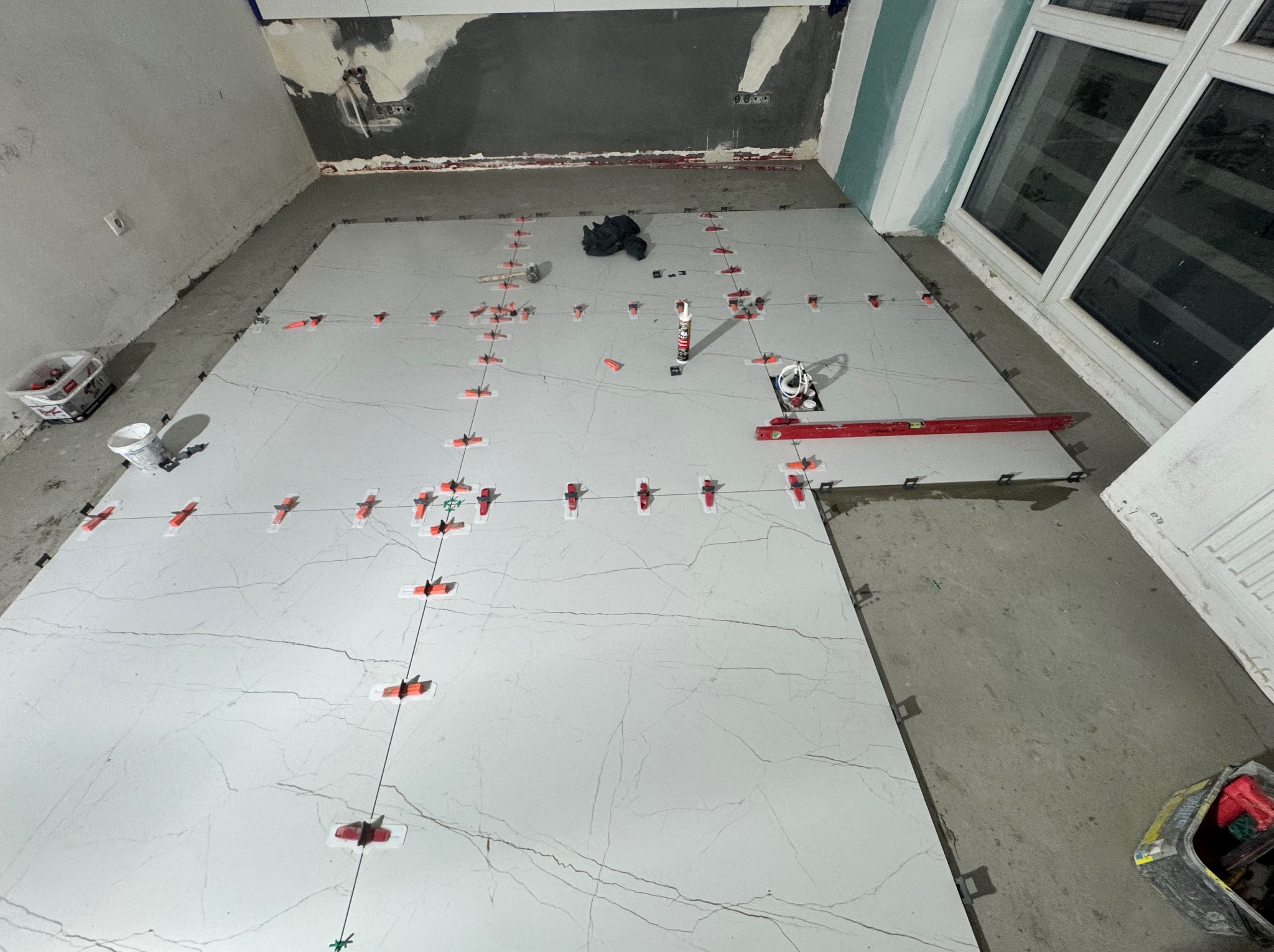 Układanie jasnych, wielkoformatowych płytek podłogowych z użyciem klipsów poziomujących. Widoczny poziomica, klej i narzędzia glazurnicze. Częściowo wykończone ściany.