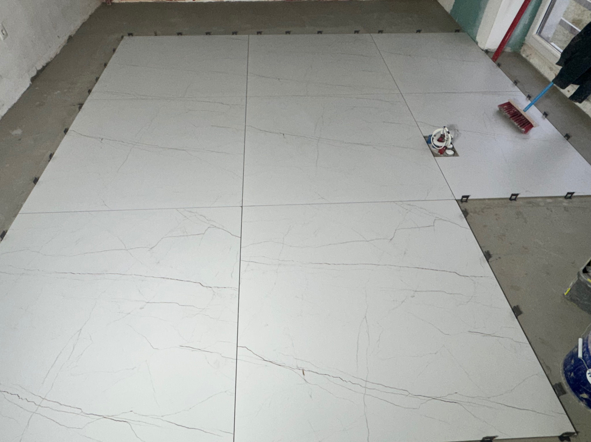 Jasne, marmurowe płytki podłogowe ułożone na podłodze z użyciem klipsów poziomujących. Widoczny fragment instalacji elektrycznej i szczotka.