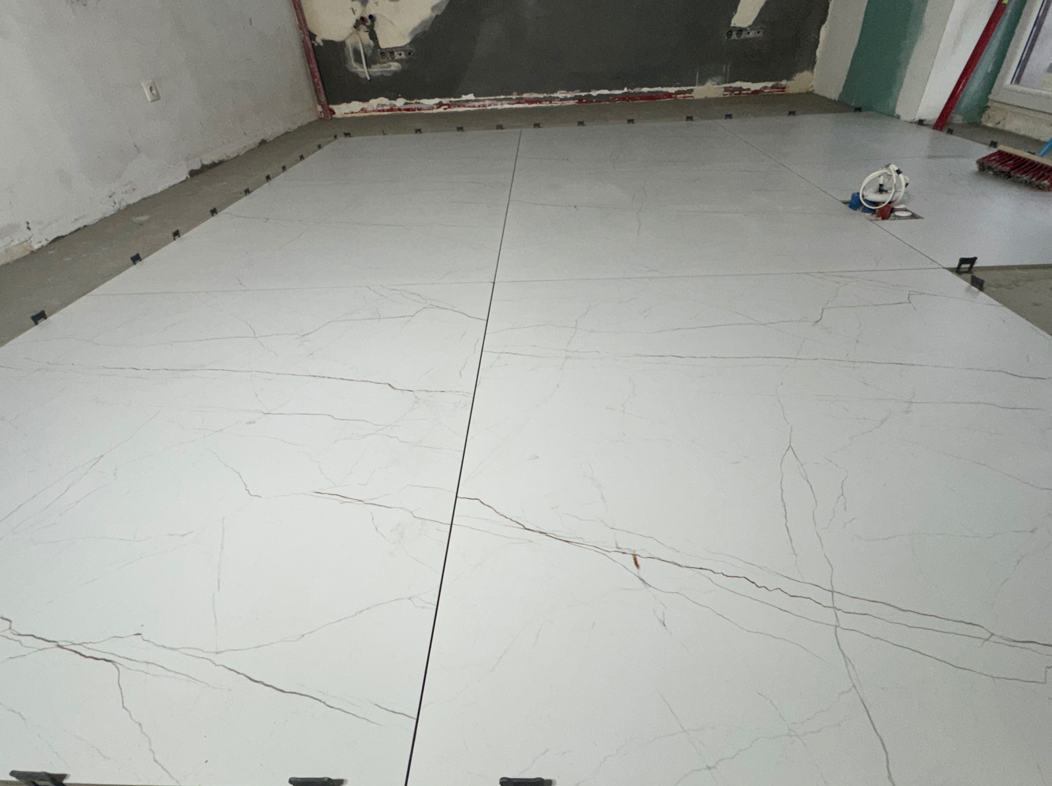 Układanie białych, wielkoformatowych płytek z szarym marmurkowym wzorem na podłodze. Widoczne czarne klipsy poziomujące i fragment ściany w trakcie prac wykończeniowych.