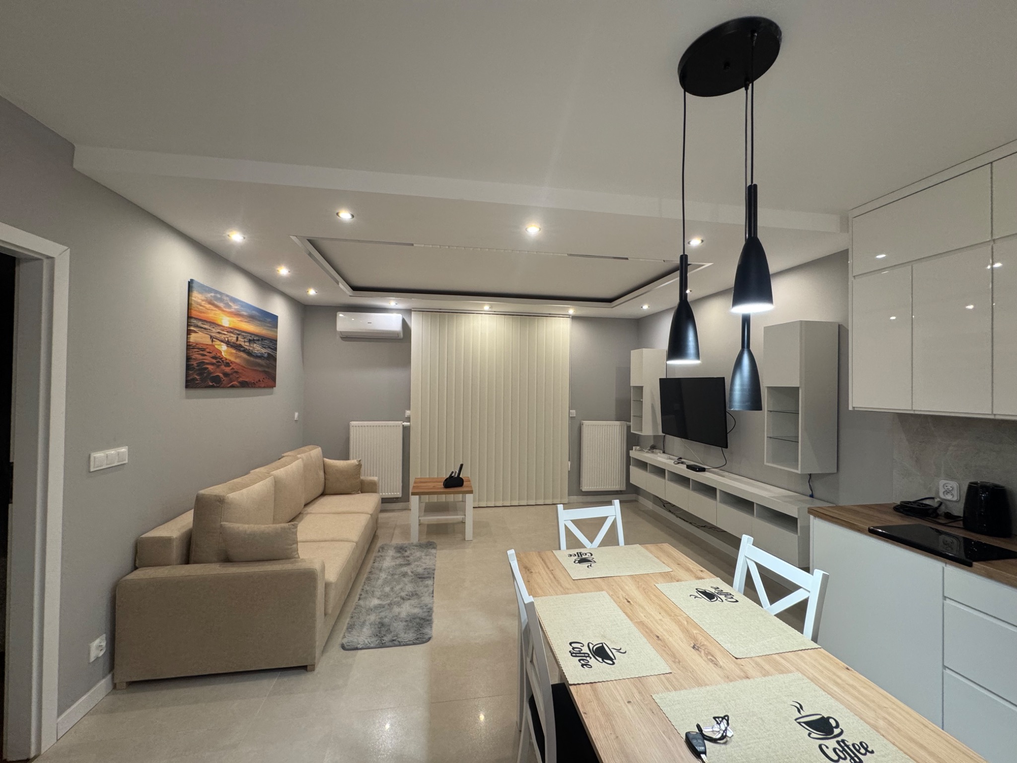 Jasne wnętrze salonu z aneksem kuchennym: beżowa sofa, drewniany stół z białymi krzesłami, białe meble kuchenne i dekoracyjne lampy wiszące. Nowoczesny design.