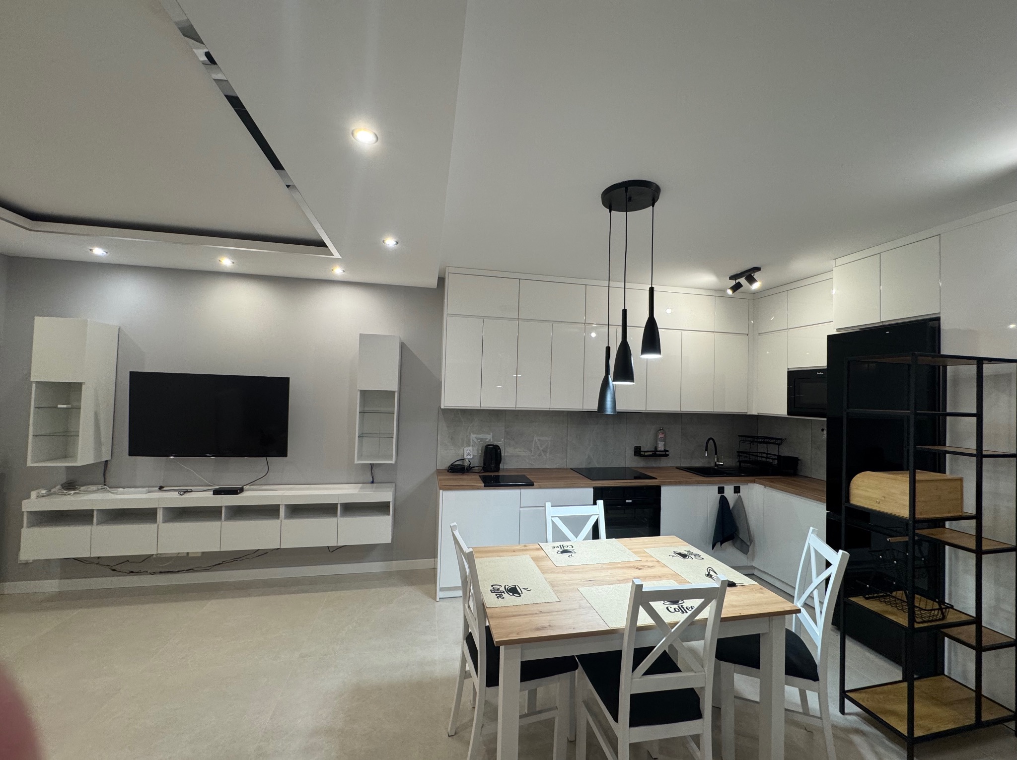 Nowoczesna, jasna kuchnia z białymi szafkami, drewnianym stołem i czarnymi lampami wiszącymi. Minimalistyczny design, szare ściany i podłoga. Uwagę przyciąga czarny regał.