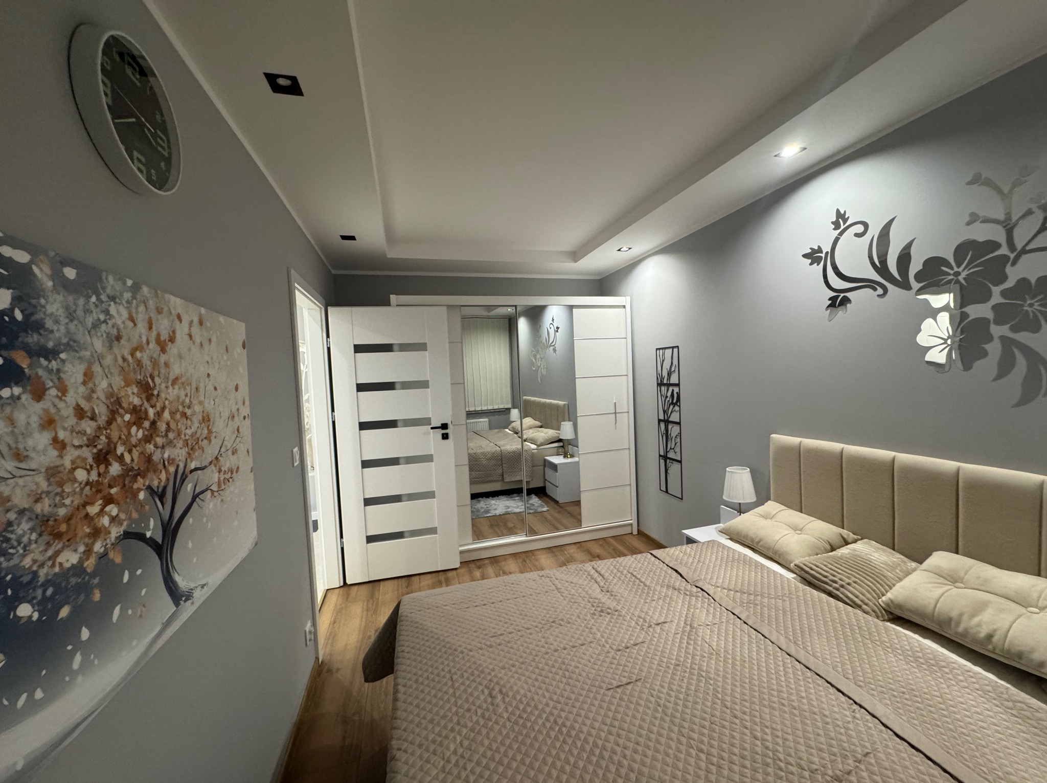 Nowoczesna sypialnia w szarościach z dużym łóżkiem, lustrzaną szafą i dekoracją ścienną w kształcie kwiatów. Widoczny obraz z motywem drzewa i zegar na ścianie.