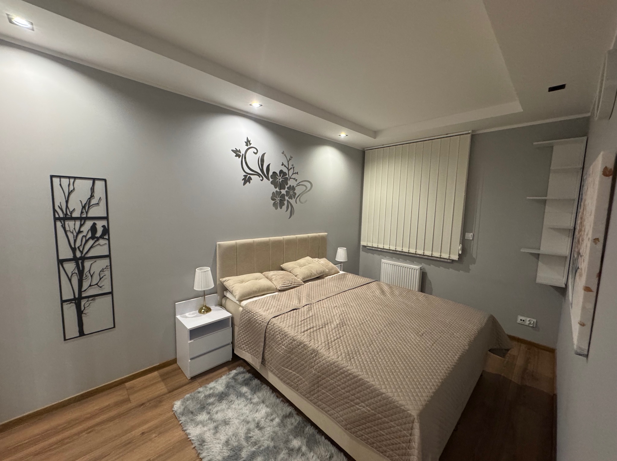 Nowoczesna sypialnia w szarościach z beżowym łóżkiem i dekoracjami ściennymi: metalowa grafika drzewa i kwiatowy motyw. Drewniana podłoga i puszysty dywan.