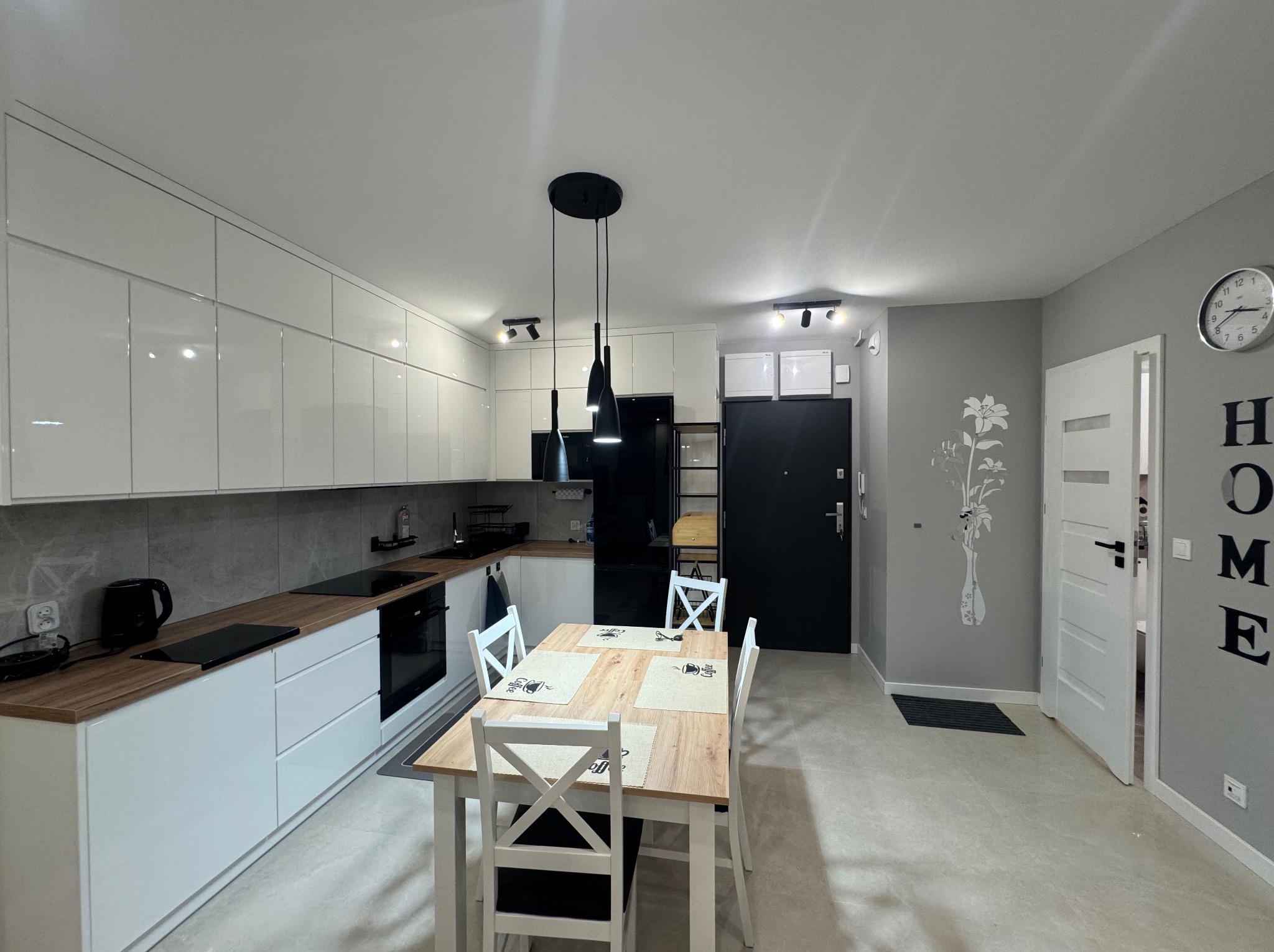 Nowoczesna kuchnia z białymi szafkami, drewnianym blatem i stołem jadalnym. Minimalistyczne oświetlenie i szara ściana z dekoracją kwiatową.