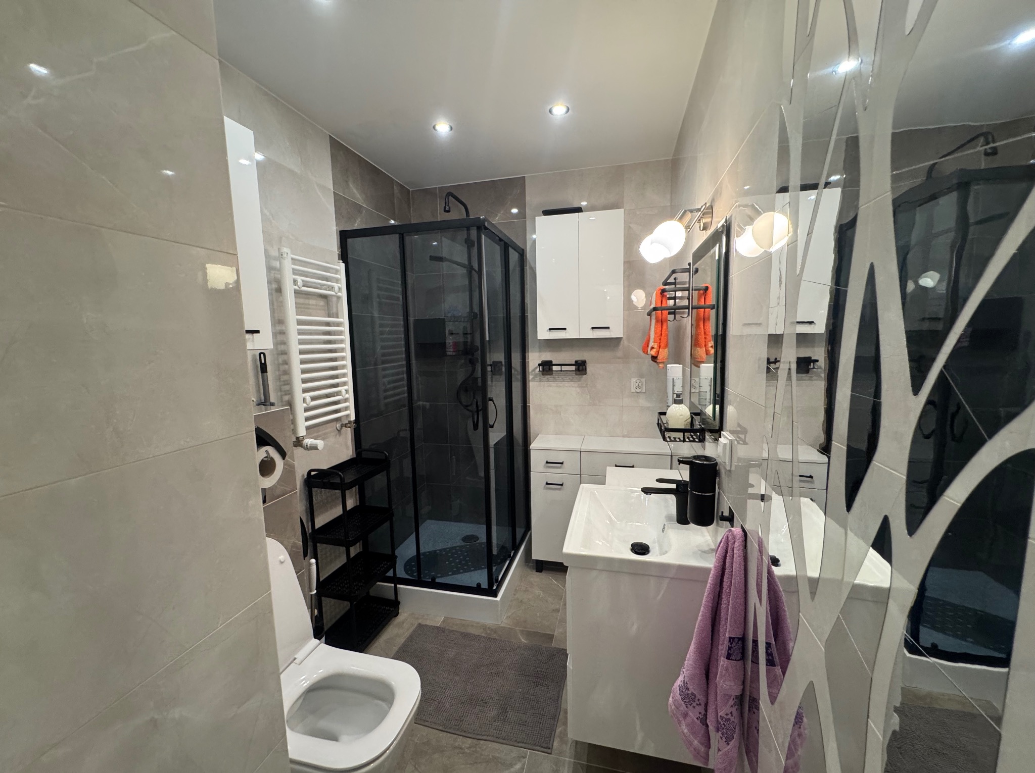 Nowoczesna łazienka w szarościach z kabiną prysznicową w czarnej ramie, białą armaturą i dekoracyjną ścianą z lustrem. Widok na całe wnętrze.