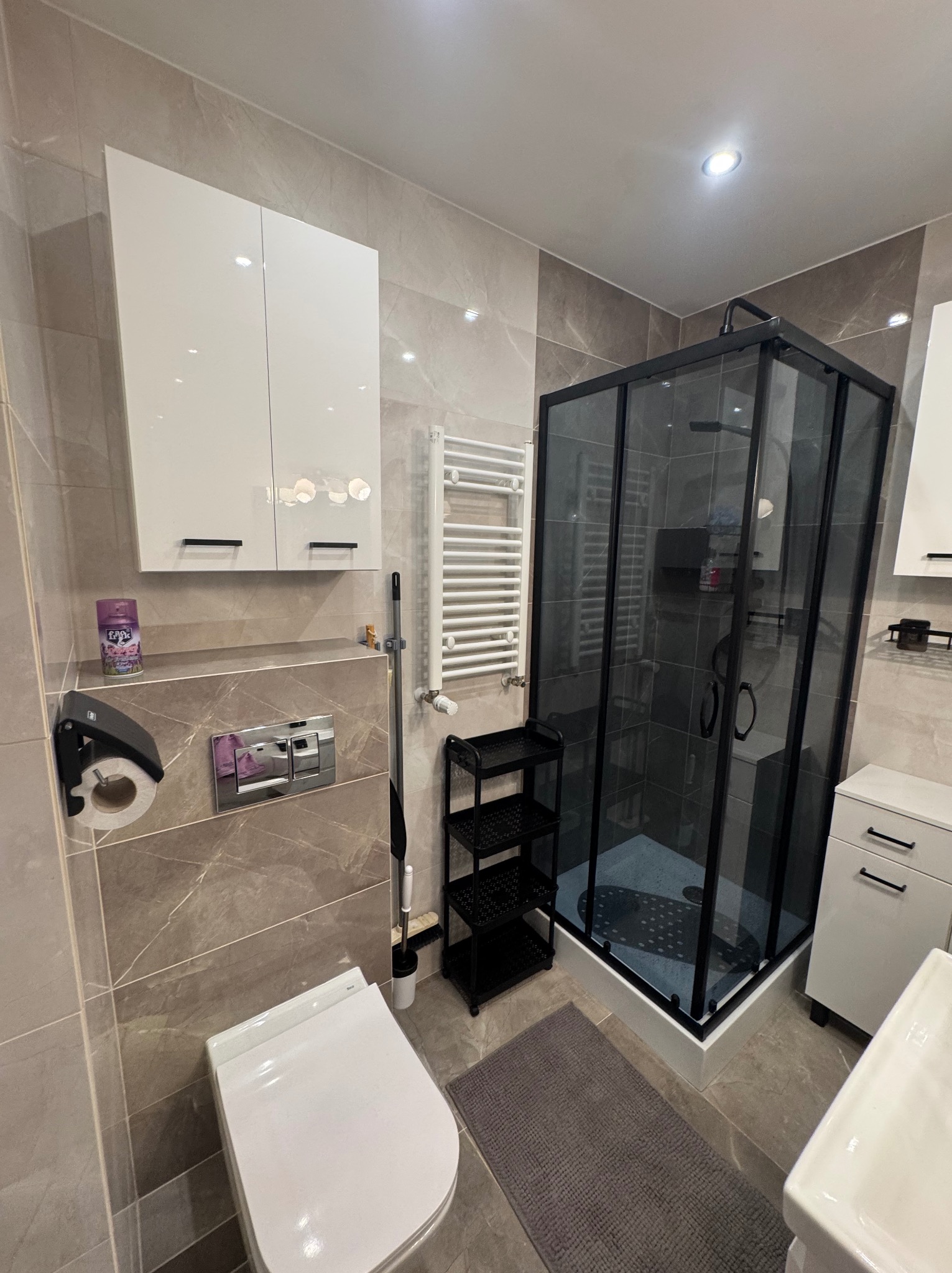 Wykończona łazienka w beżowych płytkach z kabiną prysznicową w czarnej ramie, toaletą i białymi szafkami. Grzejnik drabinkowy i czarne półki dodają funkcjonalności.