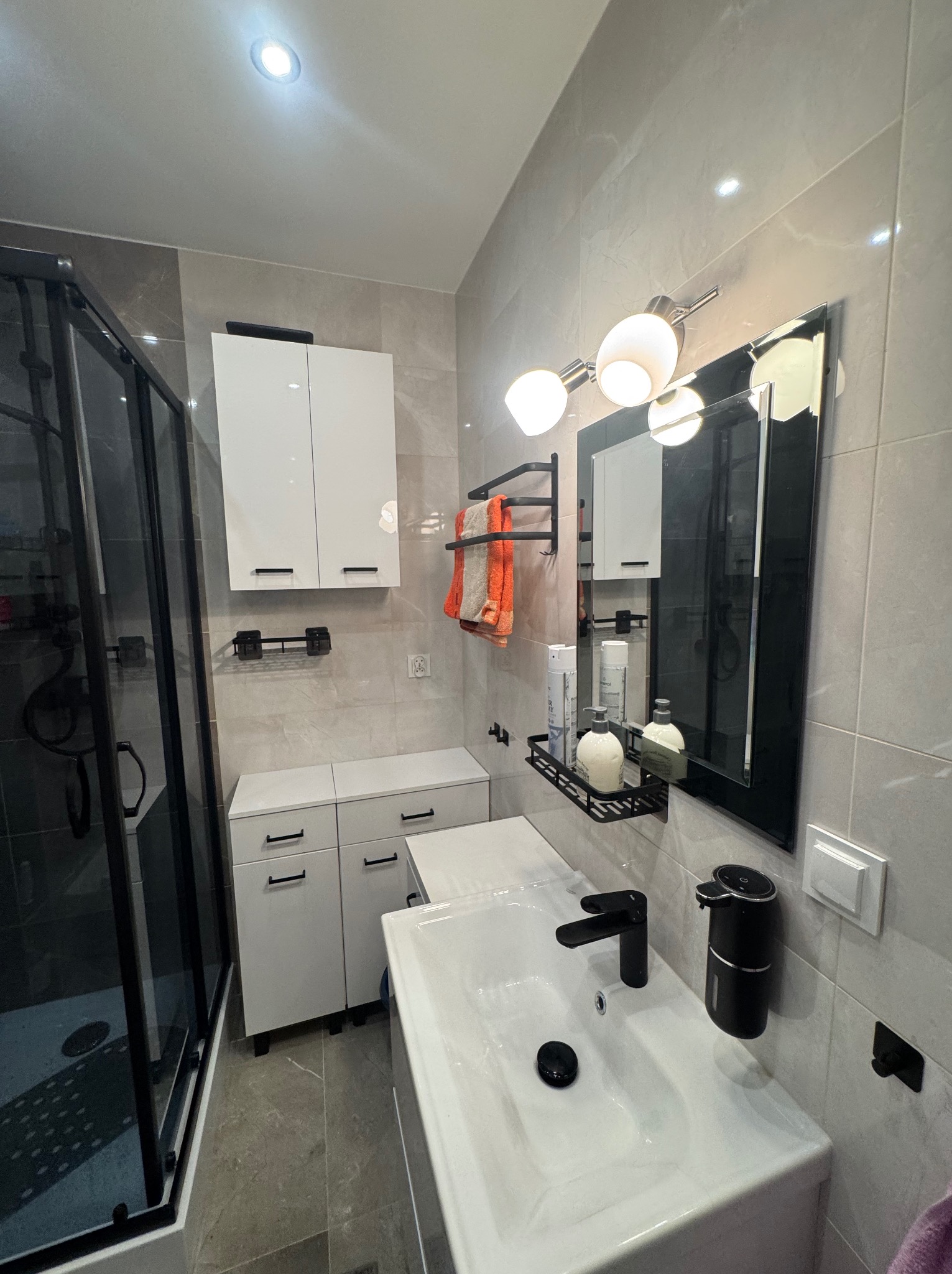 Nowoczesna łazienka po remoncie: białe szafki z czarnymi uchwytami, kabina prysznicowa z czarnymi ramami, szare płytki, czarna armatura i oświetlenie.