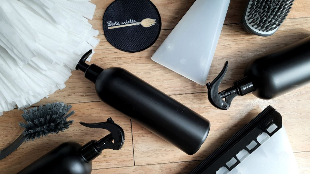 Zestaw narzędzi do sprzątania na drewnianej podłodze: mop z frędzlami, szczotki, butelki z rozpylaczami i naklejka z logo 'Złota miotła'.