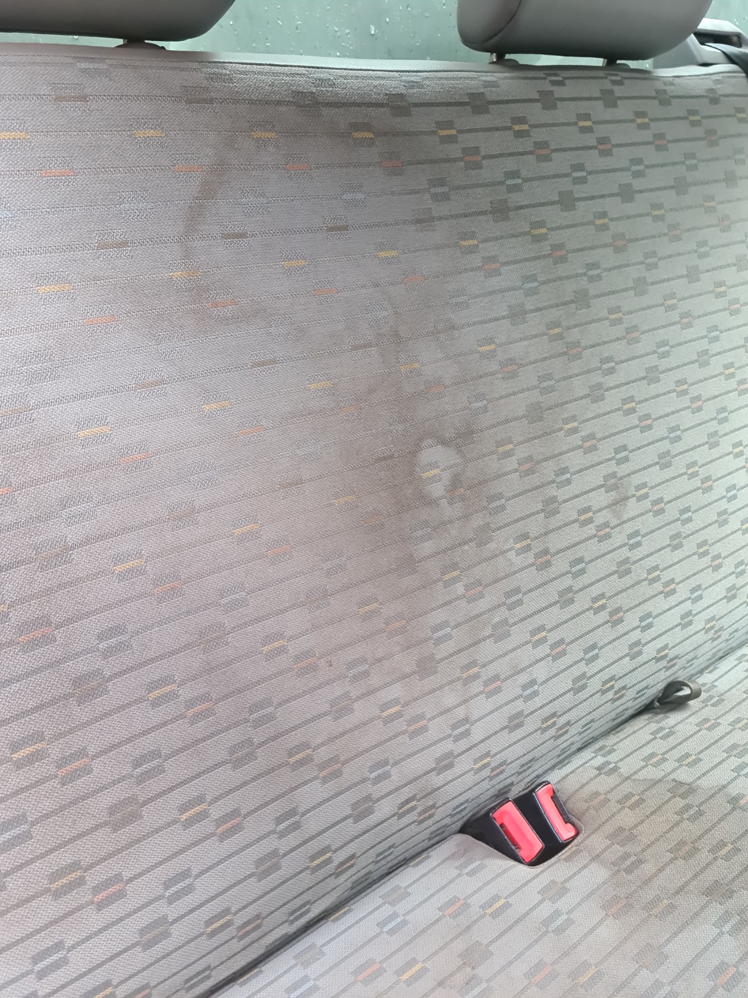 Szara tapicerka samochodowa z wzorem, widoczne plamy przed czyszczeniem, zagłówek i fragment pasów bezpieczeństwa.