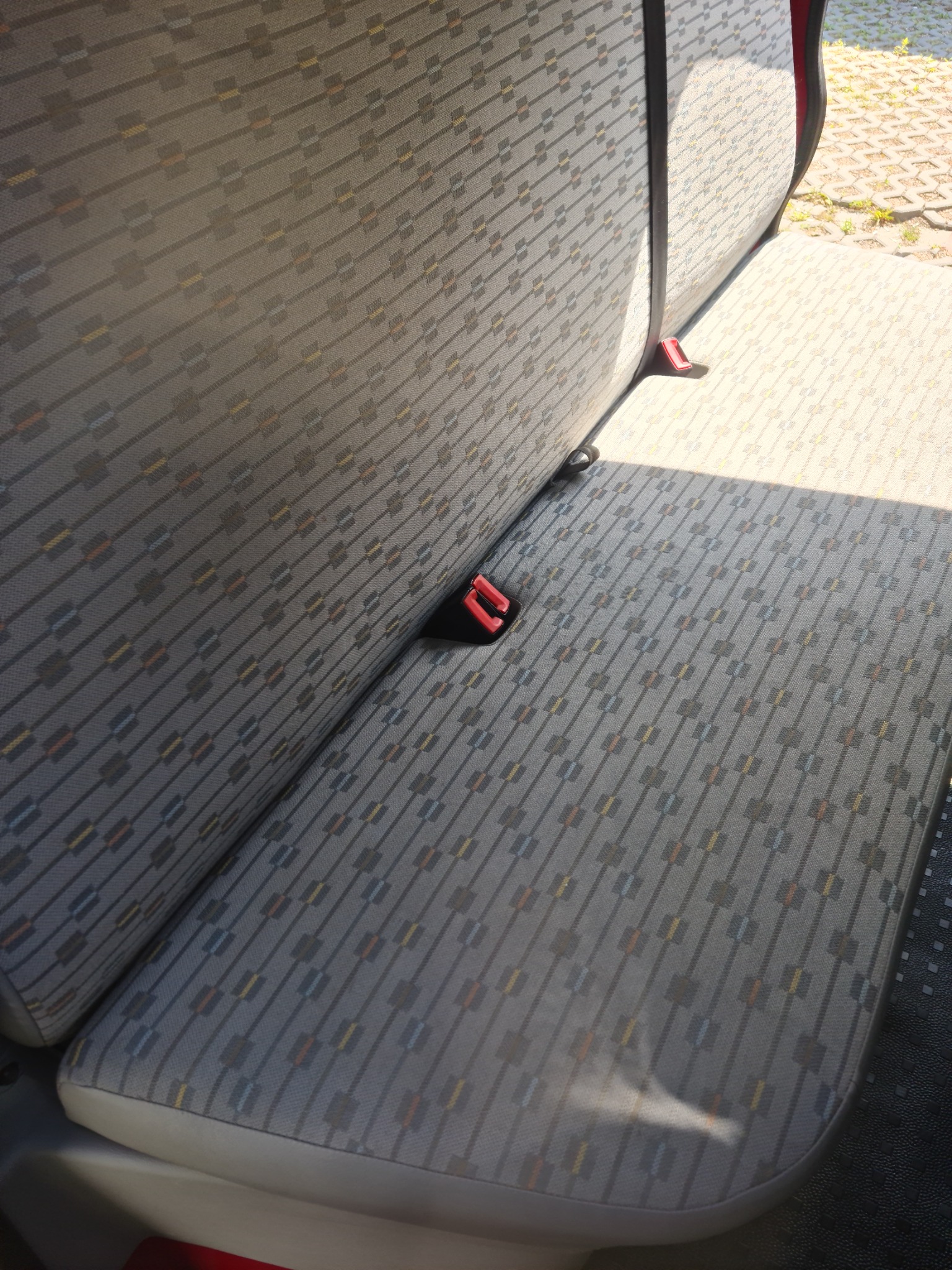 Szara tapicerka samochodowa z geometrycznym wzorem i czerwonym zapięciem pasów bezpieczeństwa, widoczne odblaski światła słonecznego.