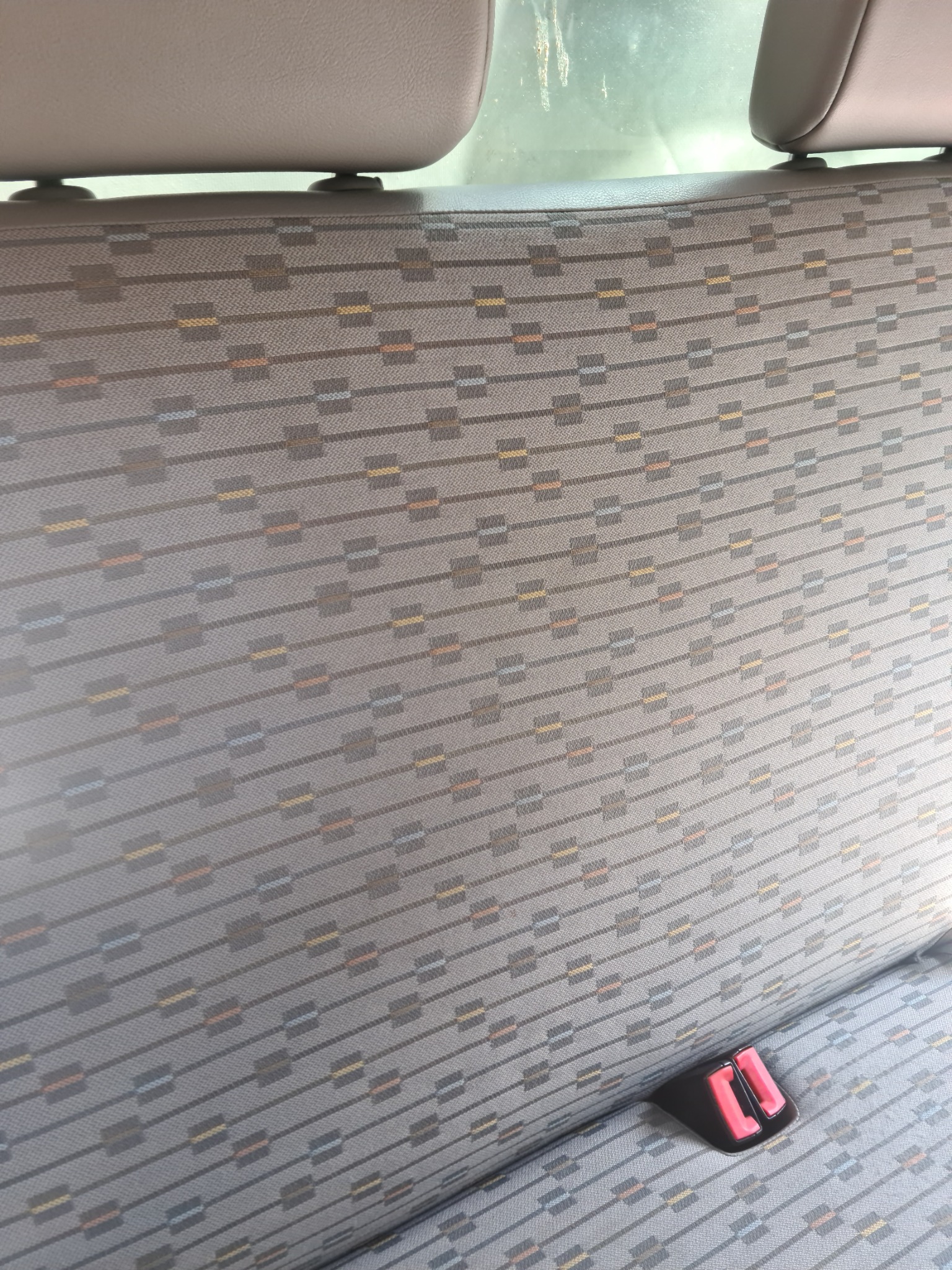 Szara tapicerka samochodowa z geometrycznym wzorem, widoczny fragment zagłówka i czerwony element zapięcia pasów bezpieczeństwa.