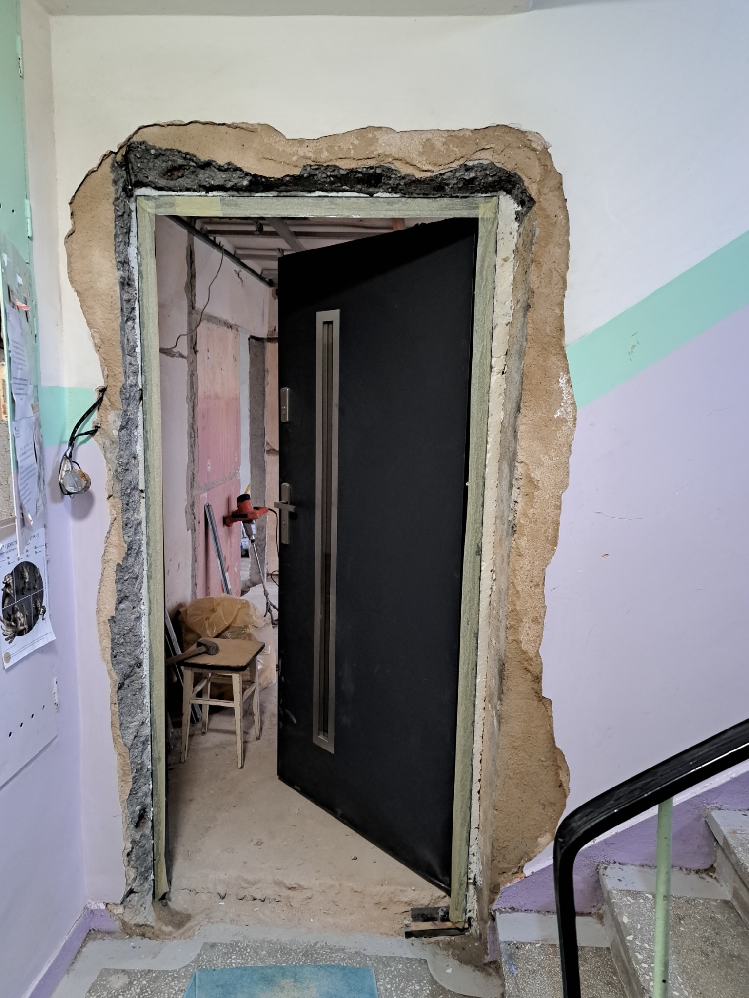 Otwarta, czarna, nowoczesna metalowa futryna drzwiowa w trakcie montażu w surowym otworze w ścianie. Widoczne ślady po kuciu, odsłonięta warstwa betonu i instalacje. W tle narzędzia i krzesło.