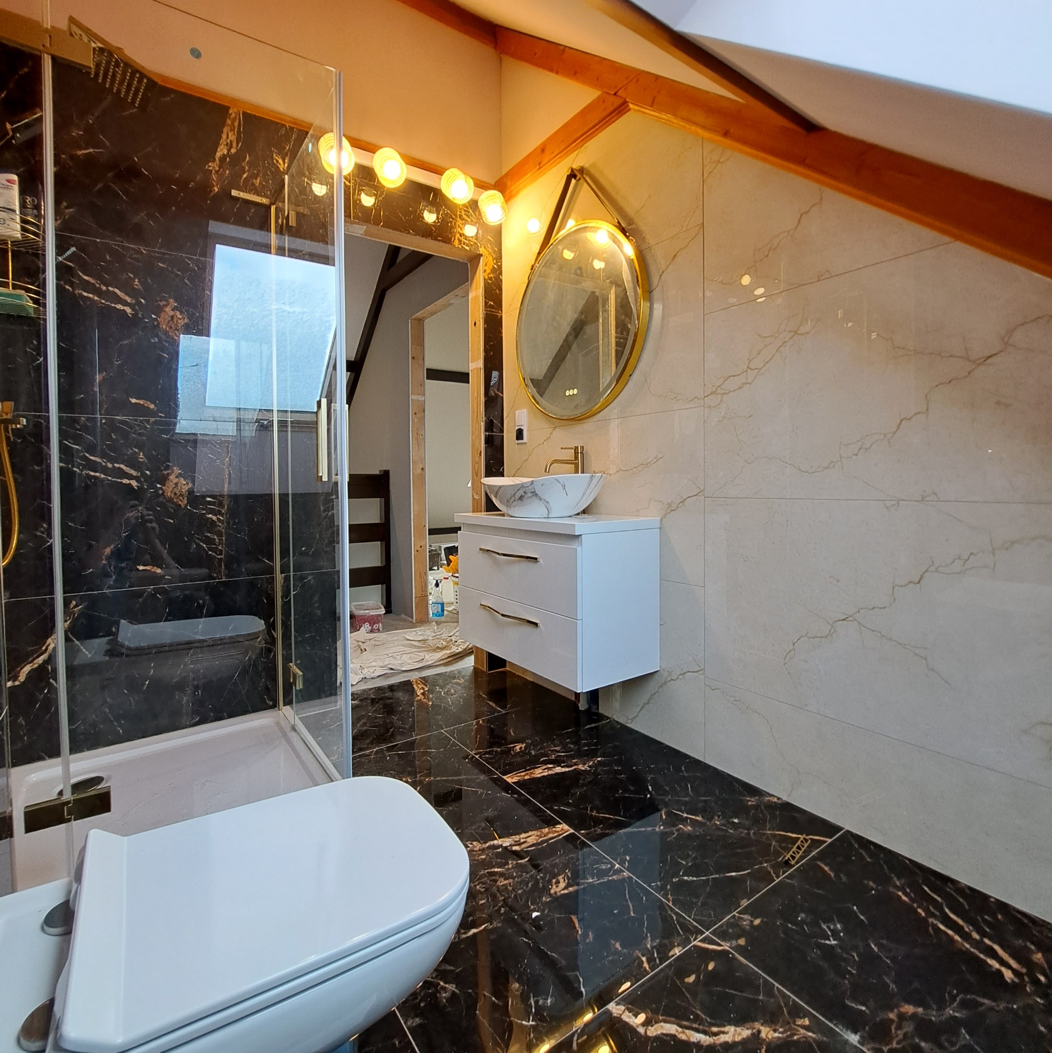 Wykończona łazienka na poddaszu z czarno-złotymi płytkami na podłodze i pod prysznicem, jasnymi płytkami na ścianach, białą szafką z umywalką nablatową, okrągłym lustrem z podświetleniem...