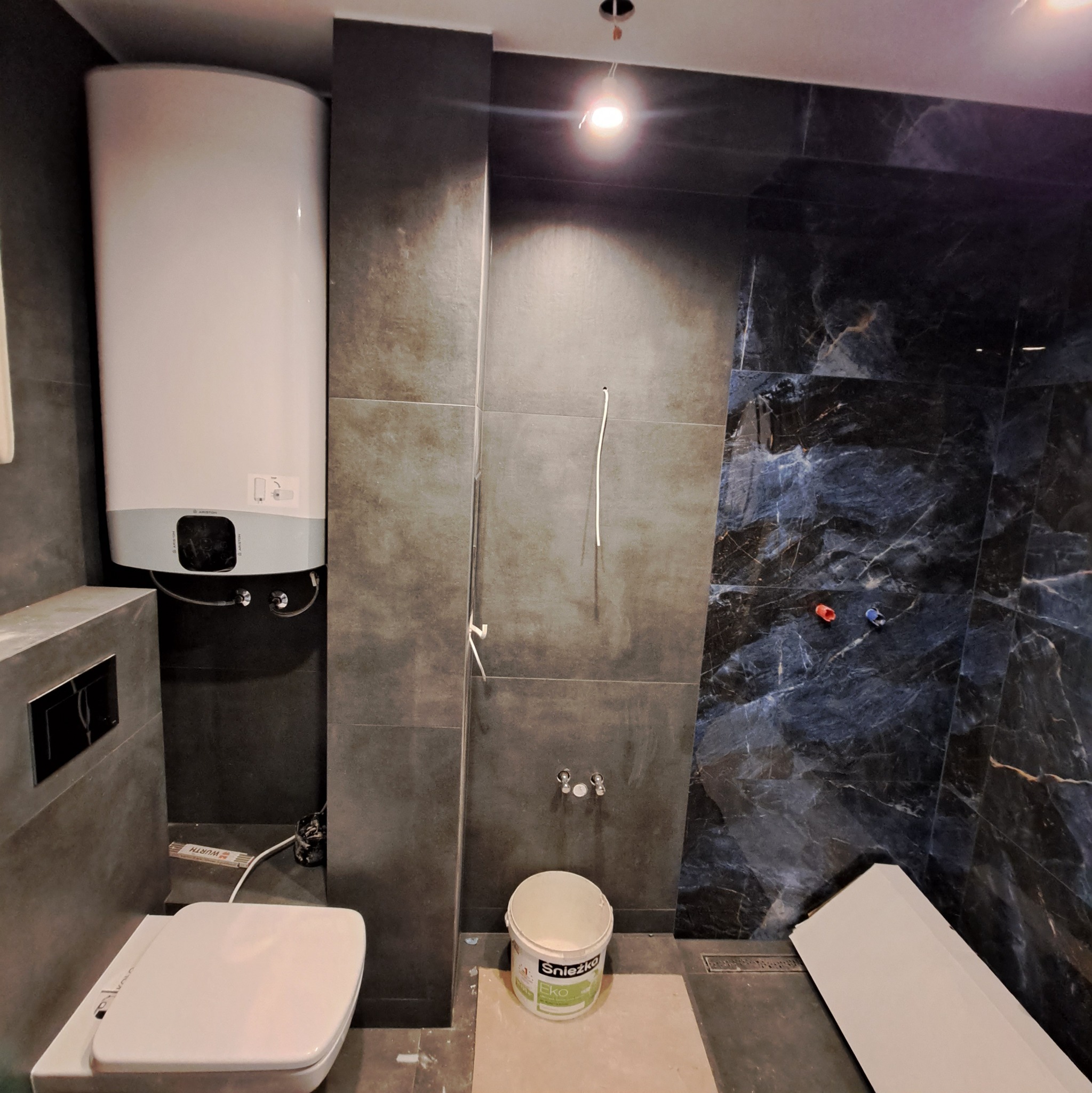 Wnętrze łazienki w trakcie prac wykończeniowych, widoczne płytki w odcieniach szarości i czerni z marmurkowym wzorem, podwieszana toaleta, bojler elektryczny oraz wiadro z farbą marki Śnieżka Eko.