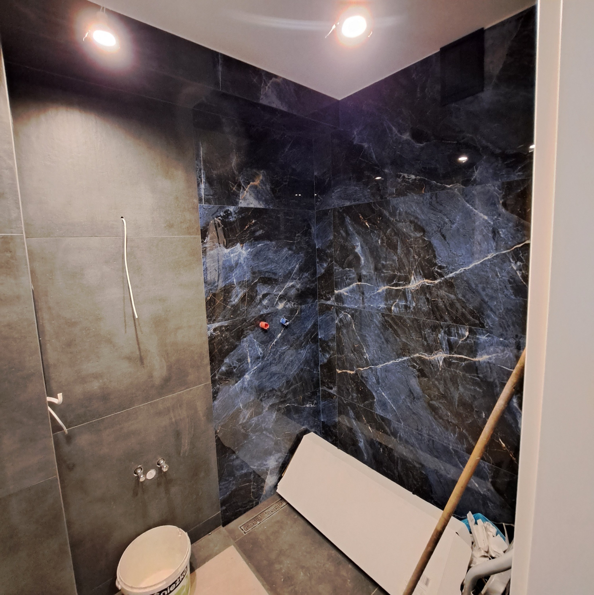 Wnętrze łazienki w trakcie remontu, z położonymi płytkami imitującymi marmur na dwóch ścianach, widoczne przyłącza wodne, wiadro z farbą i elementy wyposażenia budowlanego.