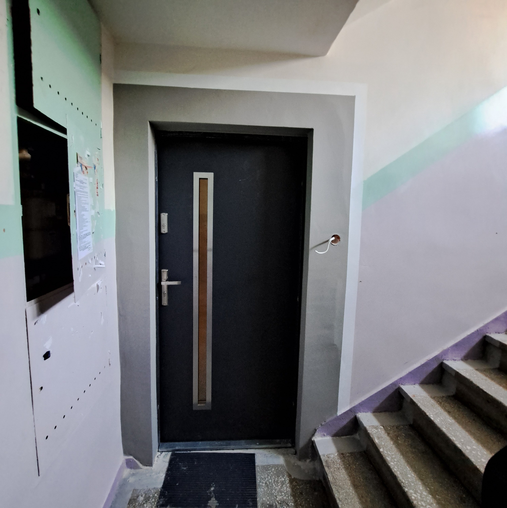 Ciemne drzwi wejściowe z metalową listwą pionową w klatce schodowej w Szczecinku, widoczne ślady po remoncie na ścianach.