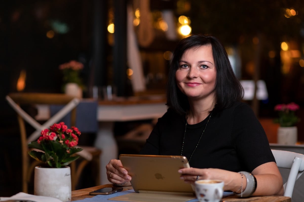 Kobieta w czarnej bluzce siedzi przy stole w kawiarni, trzyma tablet i uśmiecha się, obok filiżanka kawy i mała roślina w doniczce.