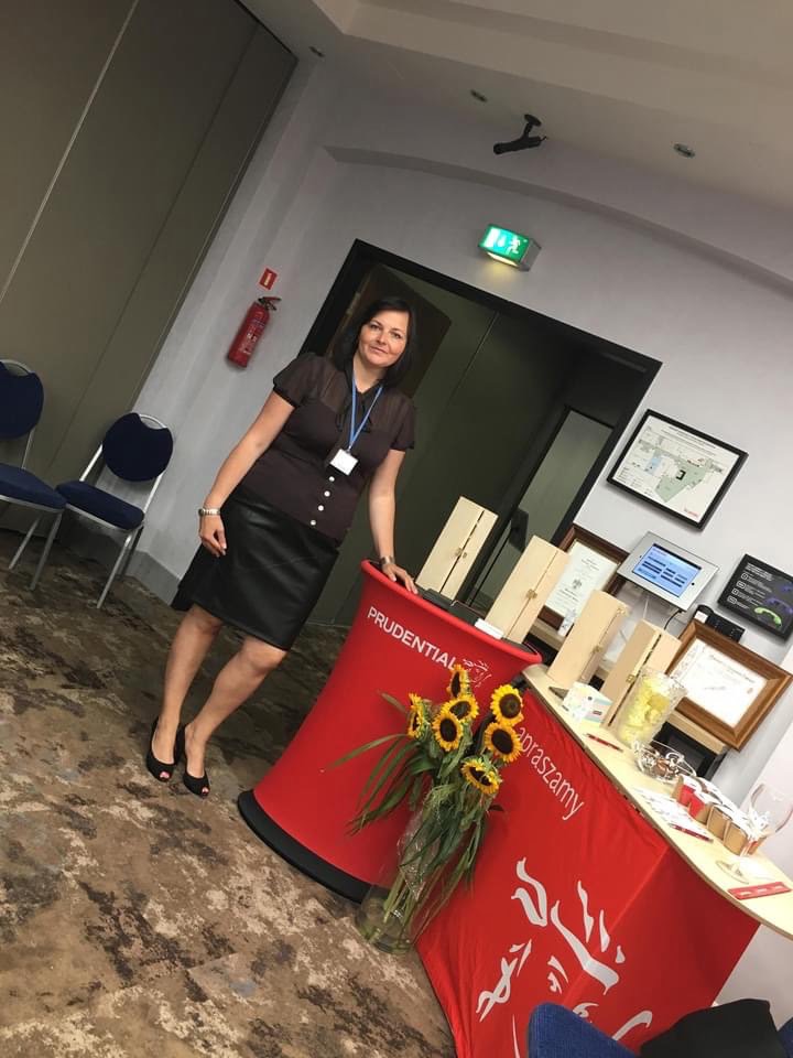 Kobieta w czarnej spódnicy i brązowej bluzce opiera się o czerwone stoisko firmy Prudential z bukietem słoneczników, na stole leżą certyfikaty i tablet.