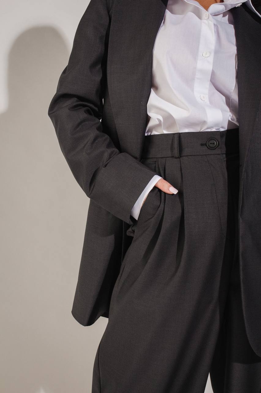 Fragment damskiego garnituru w odcieniach szarości: marynarka i spodnie z zakładkami, biała koszula z mankietami wystającymi spod rękawa marynarki, dłoń w kieszeni spodni.