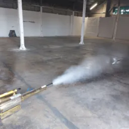 Zamgławiacz termiczny w akcji, rozpylający gęstą mgłę w pustym magazynie z betonową podłogą i białymi ścianami.