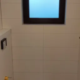 Wykończona łazienka z drewnopodobną podłogą, białymi płytkami ściennymi i oknem z mleczną szybą, widoczne elementy instalacji sanitarnej.