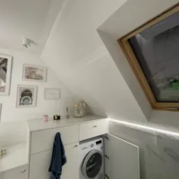 Jasna łazienka na poddaszu z pralką ukrytą w zabudowie meblowej, białe płytki imitujące marmur, okno dachowe i dekoracje ścienne tworzą przytulną przestrzeń.