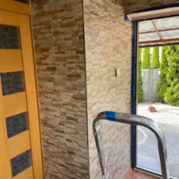 Fragment wnętrza z dekoracyjną ścianą z imitacji kamienia, widoczna aluminiowa drabina oparta o ścianę przy oknie, zabezpieczonym niebieską folią malarską.