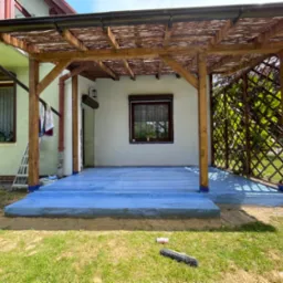 Zadaszony taras z drewnianą konstrukcją i pomalowaną na niebiesko posadzką. Widoczne zabezpieczenia folią malarską na słupach i podmurówce. Na trawie leży pędzel i pojemnik po farbie.