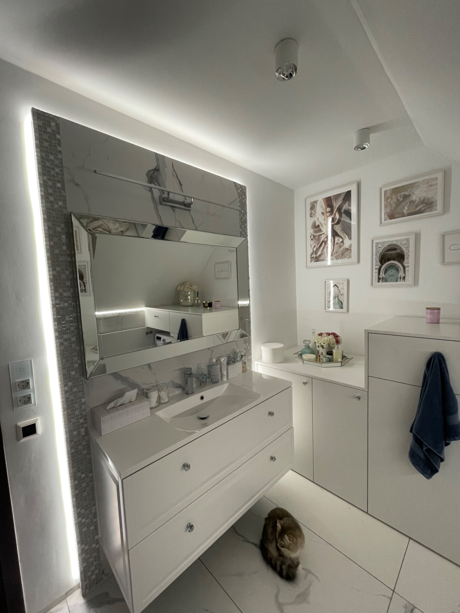 Nowoczesna łazienka z białymi meblami, lustrem z dekoracyjną ramą, oświetleniem LED i elementami marmuru; na podłodze leży zwinięty kot.