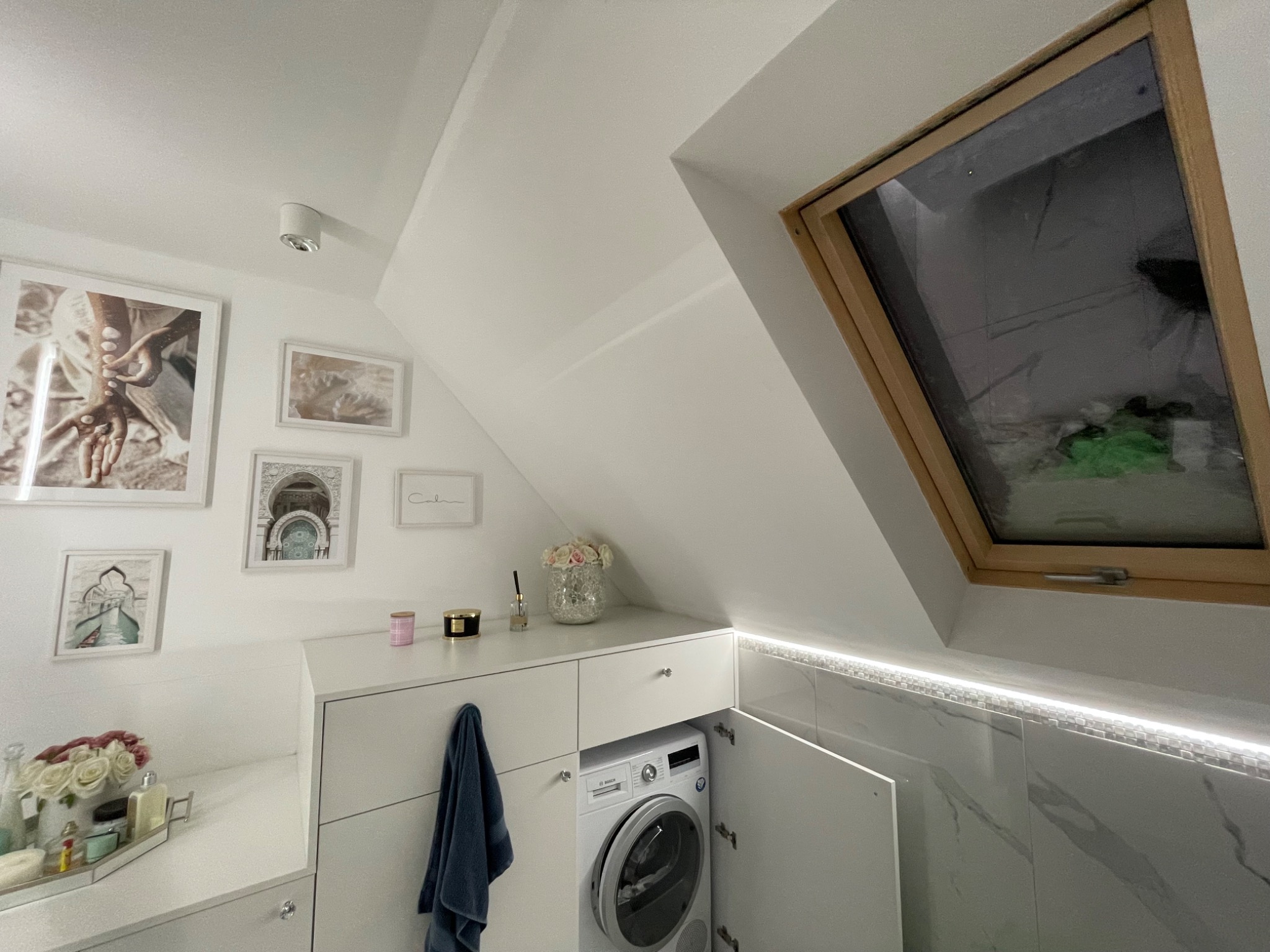 Jasna łazienka na poddaszu z pralką ukrytą w zabudowie meblowej, białe płytki imitujące marmur, okno dachowe i dekoracje ścienne tworzą przytulną przestrzeń.