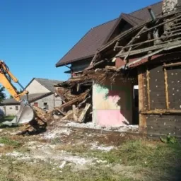 Pomarańczowa koparka w trakcie rozbiórki budynku, fragmenty dachu z ceglanym kominem, gruz i resztki konstrukcji na trawiastym terenie.