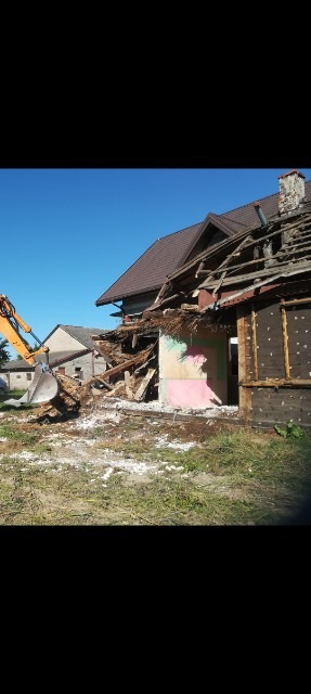 Pomarańczowa koparka w trakcie rozbiórki budynku, fragmenty dachu z ceglanym kominem, gruz i resztki konstrukcji na trawiastym terenie.