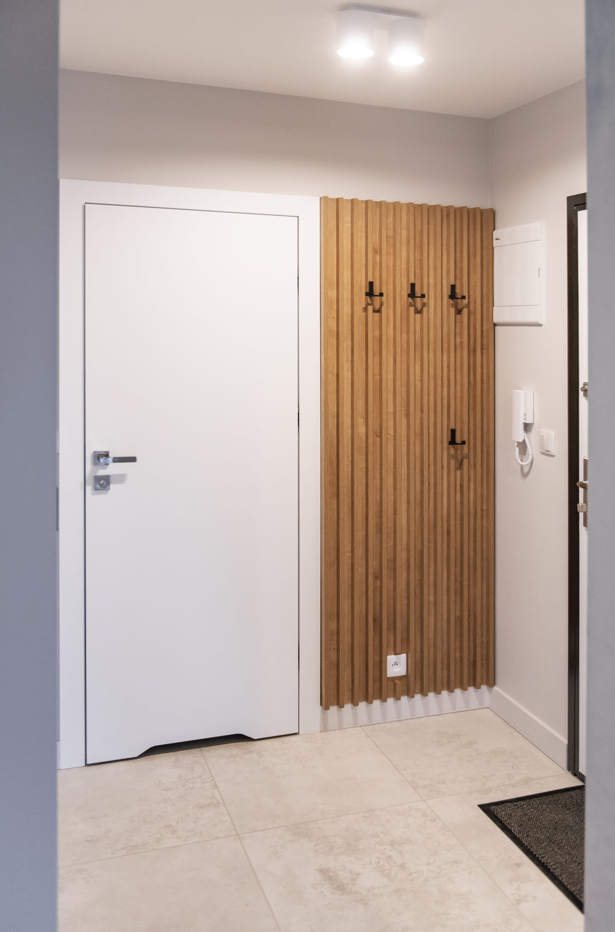 Nowoczesny przedpokój z białą, minimalistyczną szafą wnękową i panelem ściennym z drewnianych listew z wieszakami oraz gniazdkiem elektrycznym u dołu.