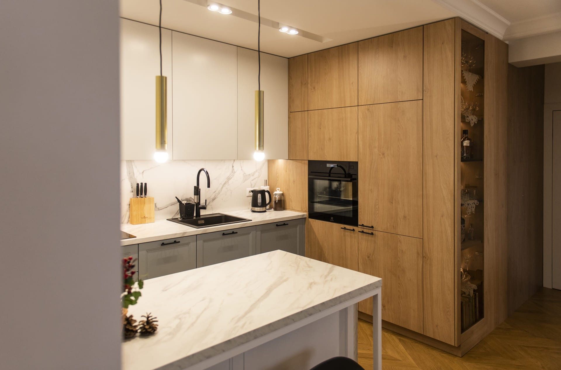 Nowoczesna kuchnia: połączenie drewnopodobnych frontów, szarości i marmuru, czarny zlew i armatura, wyspa z marmurowym blatem, oświetlenie punktowe i wiszące lampy tuby.