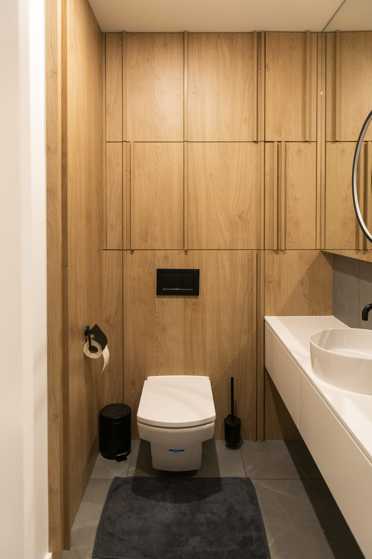 Nowoczesna łazienka z drewnianą zabudową na całej ścianie, wisząca toaleta z czarnym przyciskiem spłukującym, biała umywalka nablatowa na szafce, szara podłoga z dywanikiem.
