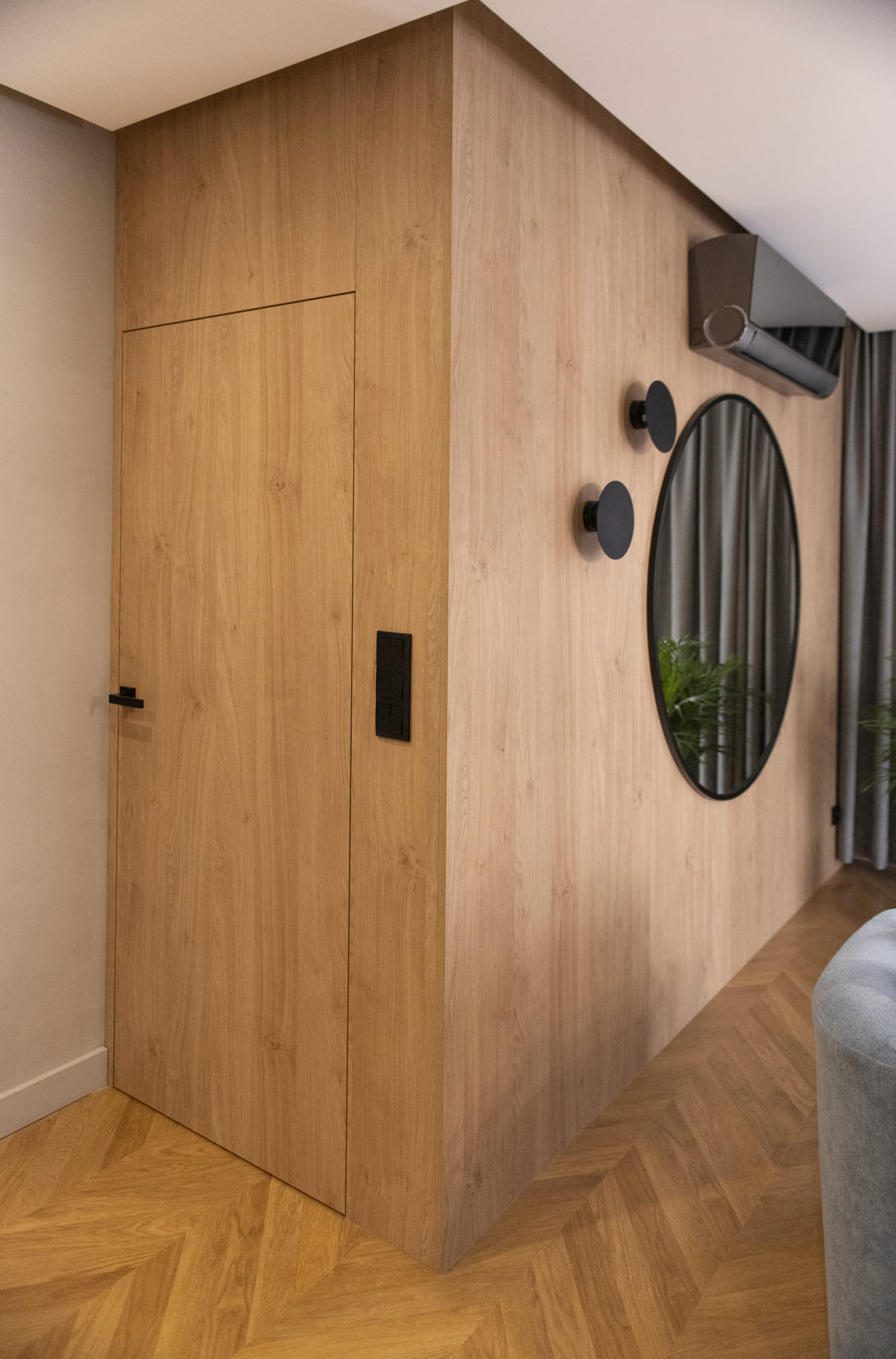 Drewniana zabudowa ściany z ukrytymi drzwiami, okrągłym lustrem w czarnej ramie, klimatyzatorem i czarnymi wieszakami na tle parkietu ułożonego w jodełkę.