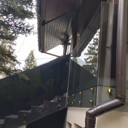 Glass4Home Adrian Kordasz - Szklana balustrada zamontowana na schodach zewnętrznych, widoczne złote mocowania i brązowa rynna, tło stanowi budynek i drzewa.
