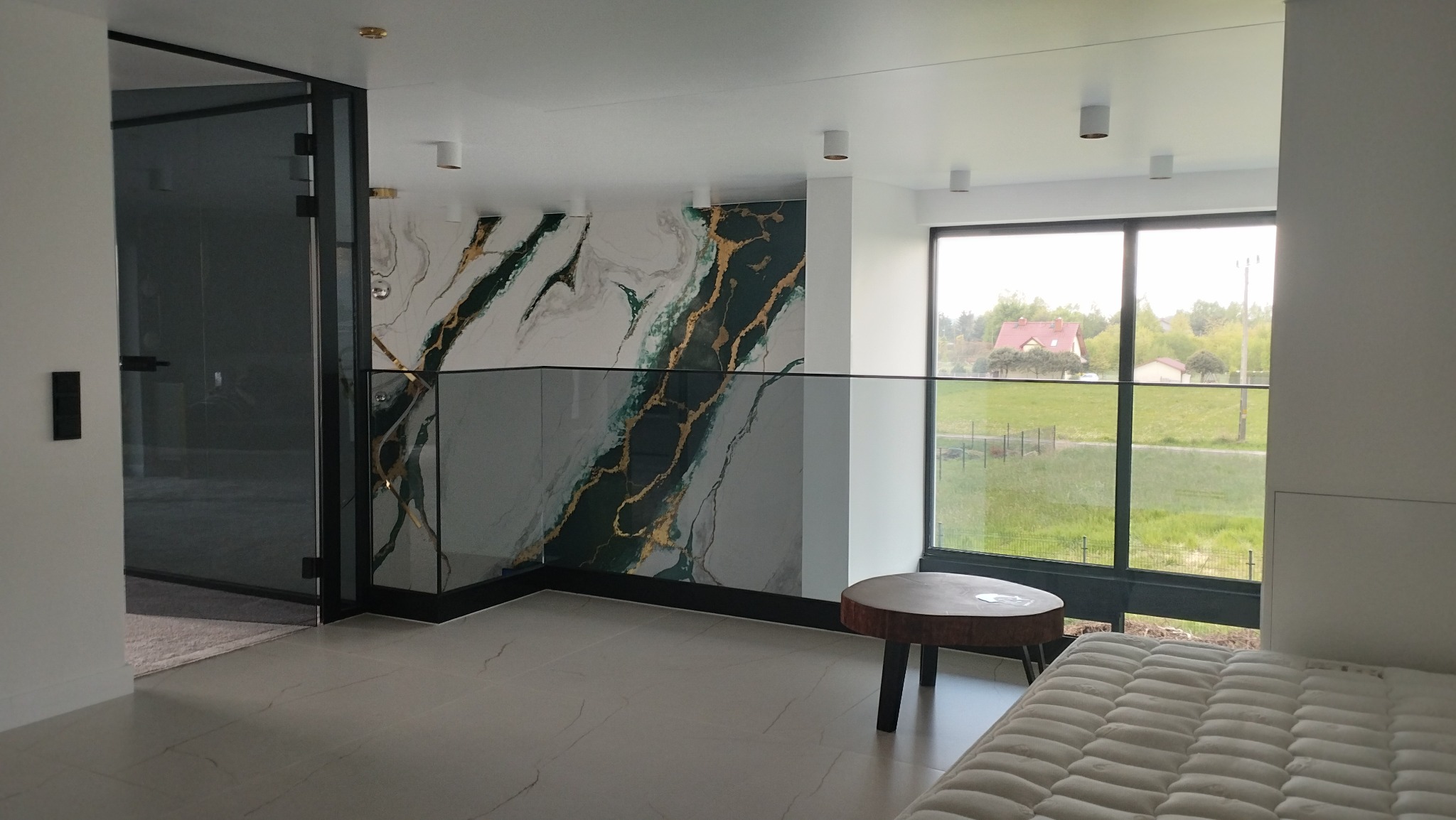 Nowoczesne wnętrze z balustradą ze szkła, marmurową ścianą z zielono-złotymi żyłami i widokiem na ogród przez duże okno, minimalistyczny stolik kawowy z drewna.