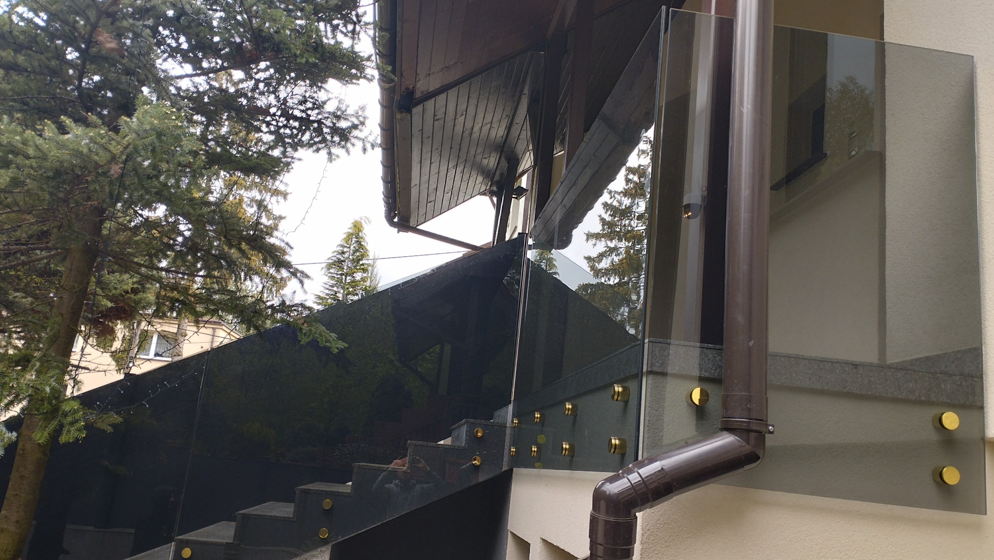 Szklana balustrada zamontowana na schodach zewnętrznych, widoczne złote mocowania i brązowa rynna, tło stanowi budynek i drzewa.