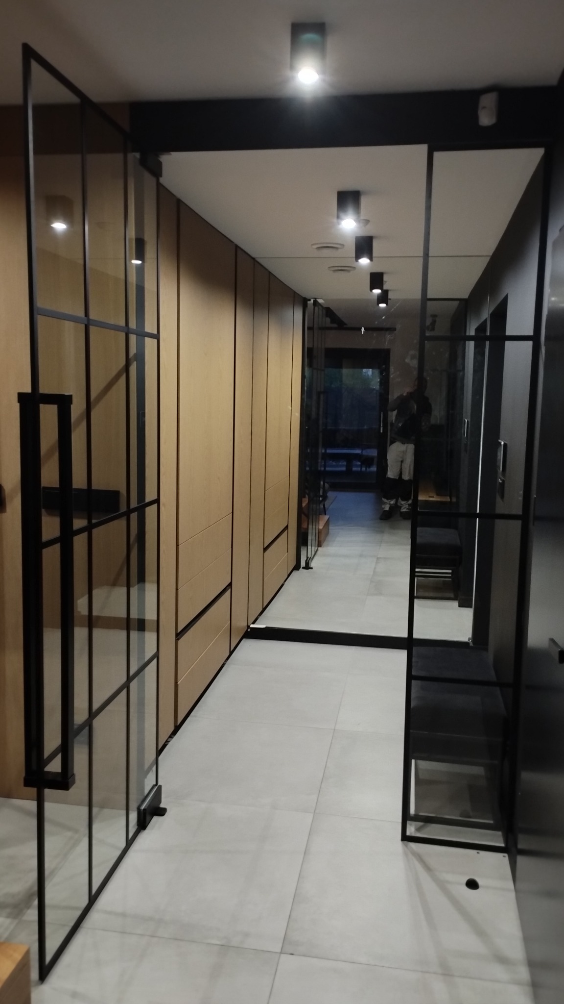 Nowoczesne wnętrze z przeszklonymi drzwiami w czarnej ramie, oddzielającymi korytarz z zabudowaną szafą w odcieniach drewna, widoczne oświetlenie sufitowe i posadzka z jasnych płytek.