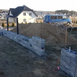 Wznoszenie ogrodzenia z szarej bloczków betonowych, widoczny fragment muru i sterta piasku na tle zabudowy jednorodzinnej oraz niebieski van.
