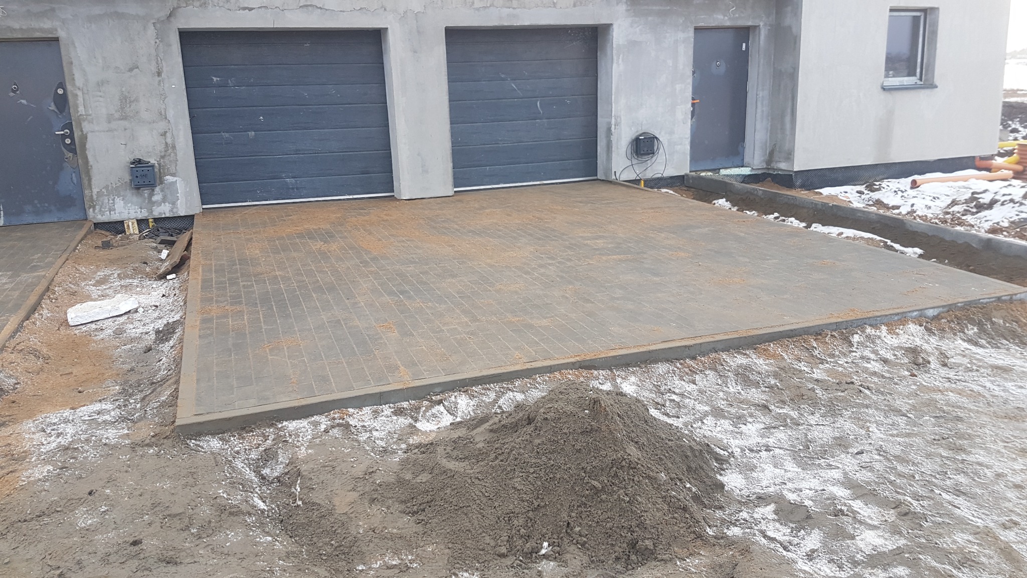 Ułożona szara kostka brukowa przed nowym domem z dwiema bramami garażowymi, widoczny piasek i resztki śniegu, etap prac wykończeniowych.