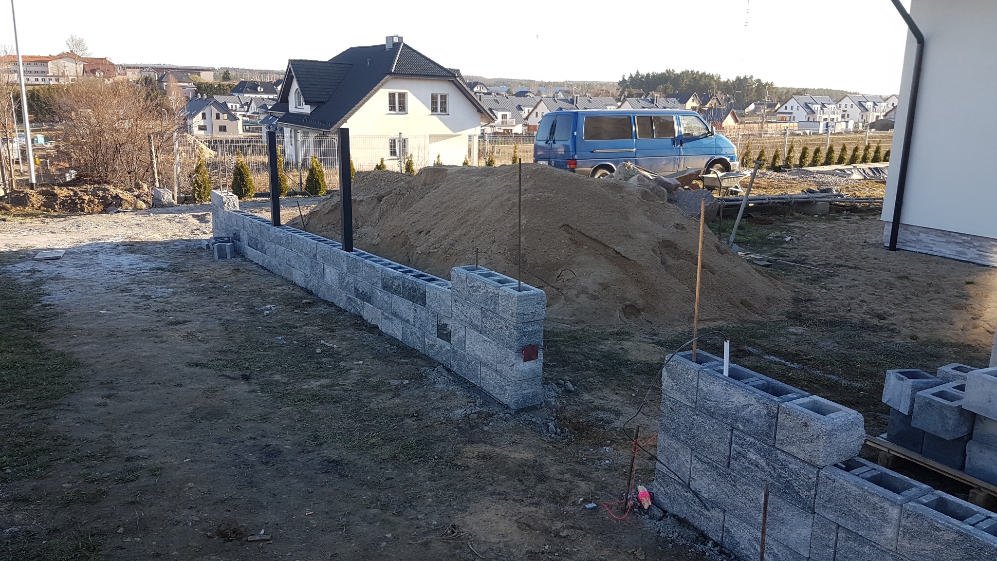Wznoszenie ogrodzenia z szarej bloczków betonowych, widoczny fragment muru i sterta piasku na tle zabudowy jednorodzinnej oraz niebieski van.