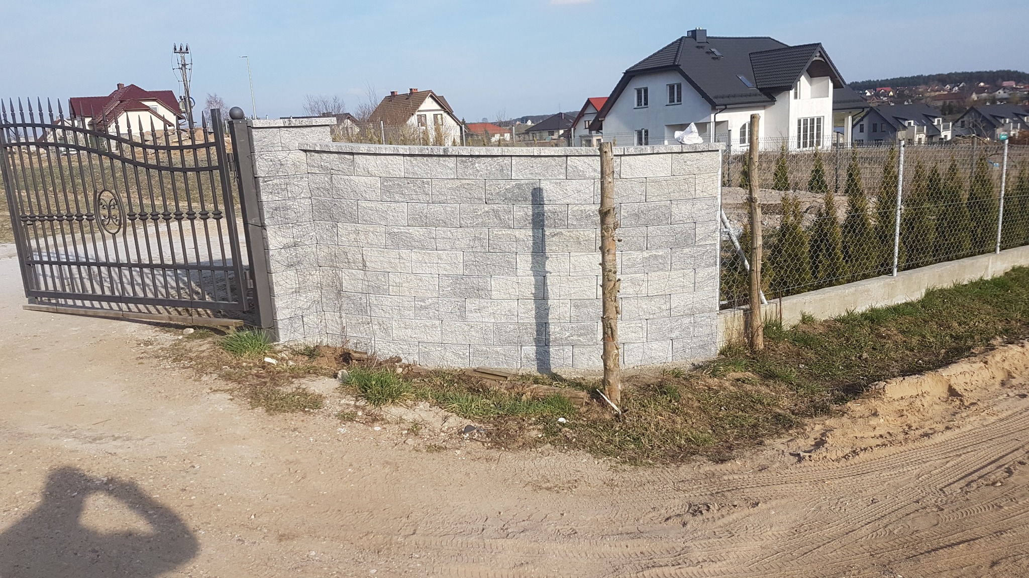 Otwarta, metalowa brama wjazdowa z ozdobnym motywem, przylegająca do niskiego, łukowatego muru z szarej kostki, a w tle widoczne domy jednorodzinne i siatka ogrodzeniowa.